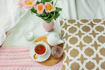 A mug of hot tea, a pink knitted blanket, a bouquet of tulips in a vase, a candle on the bed. Breakfast in bed. Cozy.