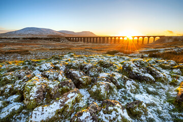 Golden light on a cold Winter evening at Ribblehead Viaduct in North Yorkshire with snow covered rocks in foreground. - obrazy, fototapety, plakaty