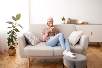 Obraz premium Full length portrait of happy young woman in hijab drinking coffee on couch at home