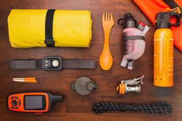 Overhead view of camping gear and equipment including headlamp, GPS, compass, rope, bear spray, water filter, and sleeping pad on a wooden background