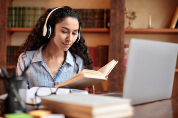 Lady sitting at desk, wearing headset, using computer, reading book - obrazy, fototapety, plakaty