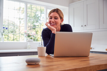 Woman In Kitchen Using Digital Smart Speaker Whilst Working From Home With Laptop During Pandemic