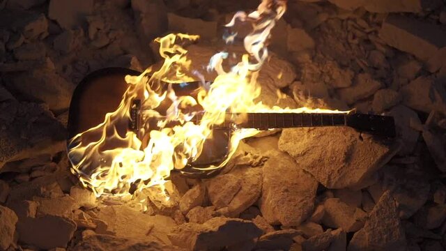 Guitar burning is on fire on stones. Acoustic western guitar burn in darkness. Flame envelopes the guitar, slow motion