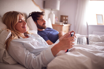 Obraz premium Couple Lying In Bed In Morning Looking At Digital Tablet And Laptop