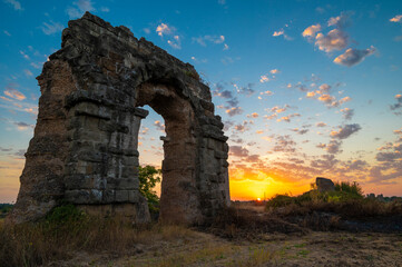 Park of the aqueducts ruins of the Claudius aqueduct, Via Appia Antica. Perspective arch during sunset, play of clouds, blue sky, yellow-orange sun reflections on the horizon. Rome, Italy. Appio latin