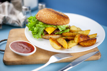 Homemade burger with potatoes and salad on a plate. Food on the table in a white plate on a wooden board. Fried potatoes and hamburger.