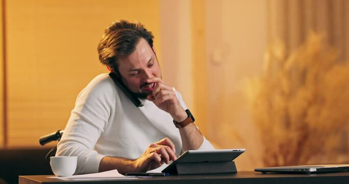 Good-looking modern purposeful young guy with well-groomed beard sitting in wheelchair after accident and distantly working on computer from his office. He is talking by phone and typing on tablet.