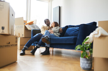 Young Couple In New Home Sitting On Sofa In Lounge On Moving Day Surrounded by Removal Boxes
