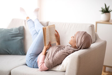Fototapeta na wymiar Cozy stay at home activities. Black young woman in hijab reading her favorite book, lying on sofa in living room