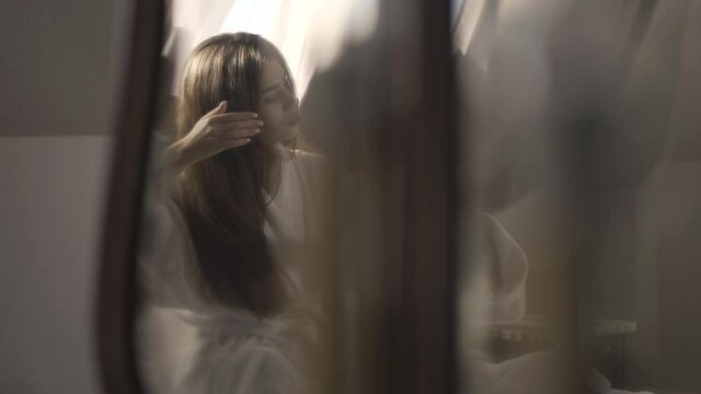 Reflection in mirror of gorgeous slim rich woman combing hair in the morning in sunny bedroom. Confident beautiful brunette Caucasian lady in white bathrobe taking care of beauty indoors at home.
