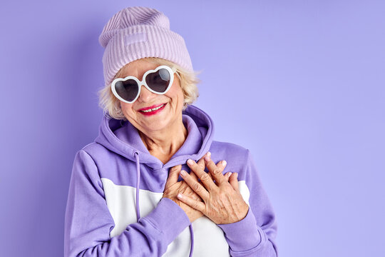 Elderly Woman Holding Hands On Chest Smiles At Camera Isolated Over Purple Background, Fancy Female In Pullover, Hat And Sunglasses