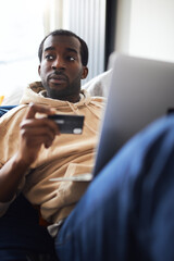 Young Man At Home Lying On Sofa With Laptop Using Credit Card To Shop Online