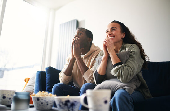 Excited Young Couple Relaxing At Home Sitting On Sofa Whilst Watching Sport On TV Together