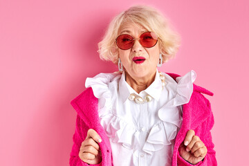 amazed elderly female in stylish clothes posing at camera isolated on pink background, surprised...