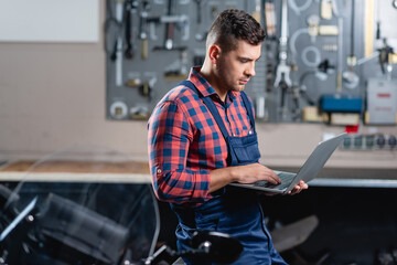 young technician in overalls using laptop in workshop, blurred foreground