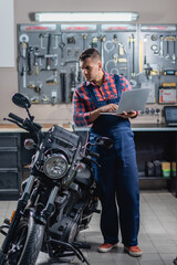 full length view of mechanic in overalls using laptop near motorbike in garage