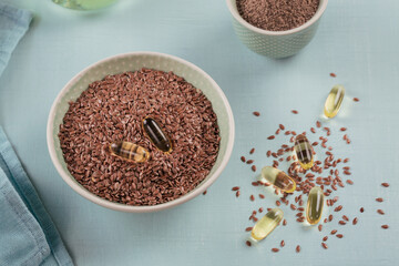 Brown flax seed or linseed and ground or crushed flaxseed in small bowl and gelatin capsules with omega oil on a light blue background.