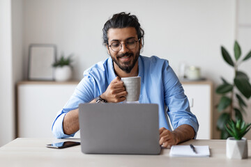Young Smiling Arab Freelancer Guy Using Laptop And Drinking Coffee At Home
