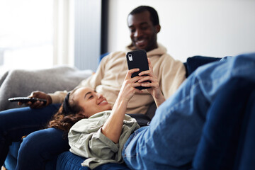 Relaxed Young Couple At Home Sitting On Sofa Watching TV And Checking Social Media On Mobile Phone