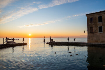 Scenic sunset at Punta San Vigilio, popular Italian touristic destination on Garda Lake, Lombardy. Clouds in the sky reflect in the water, people silhouette enjoying the sunset