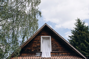 Wooden old unpainted house with a triangular sloping roof, next to the trees. Upper floor with open window, outdoors white day curtain floating in the wind. 
Draught, lightness and careless feeling