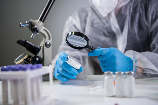 Scientist In Hazmat Suit And Goggles Holding Vaccine Bottle And Magnifying Glass, Looking And Research. Epidemiology Virus Test. Pharmaceutical Research, Clinical Trials. Biotechnology Chemist In Lab