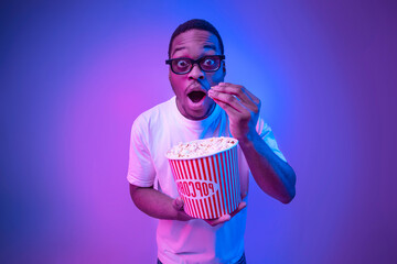 Shocked black guy in 3D glasses eating popcorn in cinema, neon light - obrazy, fototapety, plakaty