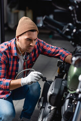 Fototapeta na wymiar mechanic in plaid shirt and gloves checking engine of motorcycle with stethoscope on blurred foreground