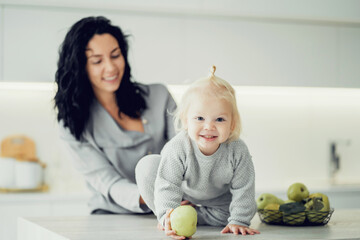 Fototapeta premium the child keeps playing with a green Apple. smiling at the camera in comfortable children's clothes in gray. young family mom and daughter at home in the kitchen.