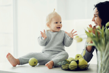 Fototapeta premium young family mother and daughter sit at home in the kitchen, the child plays with a green Apple and smiles at the camera in comfortable stylish gray clothes