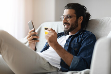 Cheerful Arab Guy Shopping Online With Smartphone And Credit Card At Home