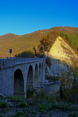  old stone bridge over the mountain river across autumn forest, hills, and sunset sky. Natural background. Travel, movement, architecture concept