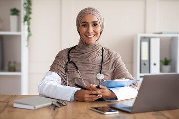 Smiling young woman doctor in hijab taking anamnesis in clinic - obrazy, fototapety, plakaty
