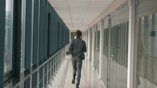 Young businessman running along long office corridor with window corridor back view of camera. Man in business suit clothing running room hall to meeting work indoors. Office style building success