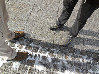 Vier Beine und Stiefel von 2 Geschäftsleuten, die auf grauem Kopfsteinpflaster auf dem Bürgersteig stehen. Die Sonne macht lange Schatten auf dem steinigen Boden. Diese Szene stammt aus Chemnitz