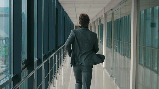Young businessman running along long office corridor with window corridor back view of camera. Man in business suit clothing running room hall to meeting work indoors. Office style building success