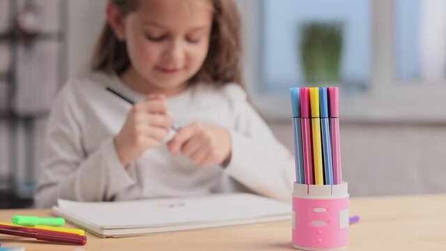 Cute little girl are drawing pictures with felt-tip pen on the white paper album sitting at the table. Playing alone, creative art activity at home. Spending free time at home during school holidays