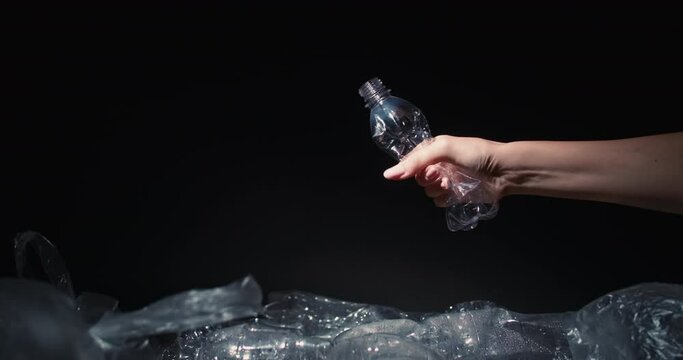 Waste reduction. Plastic recycling. Environmental protection. Female volunteer hand crushing used empty bottle throwing into trash dump with cellophane bags isolated on black copy space background.