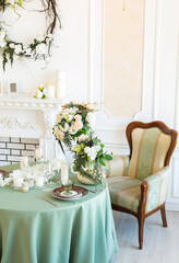 Luxury stylish bright light interior of sitting room. White walls decorated by ornament. Fireplace. Nobody inside room. Table setting by dishes, candles and flower bouquets.