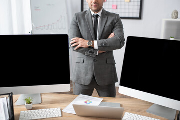 Naklejka premium cropped view of trader standing with crossed arms near laptop and computer monitors