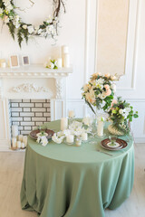 Luxury stylish bright light interior of sitting room. White walls decorated by ornament. Fireplace. Nobody inside room. Table setting by dishes, candles and flower bouquets.