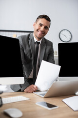 smiling trader looking at camera near computer monitors and laptop on blurred foreground