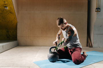 Focused young armless man training, sitting on his legs on a mat next to a kettlebell. Attaching rope support to a stump to lift weight with it.