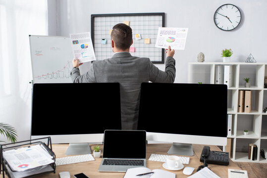 back view of trader holding infographics near monitors with blank screen on blurred foreground - Powered by Adobe