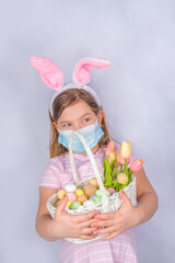 Coronavirus and Easter holiday. Cute little girl in a protective medical mask with bunny ears. Easter banner background.  with Easter eggs and spring flower in basket