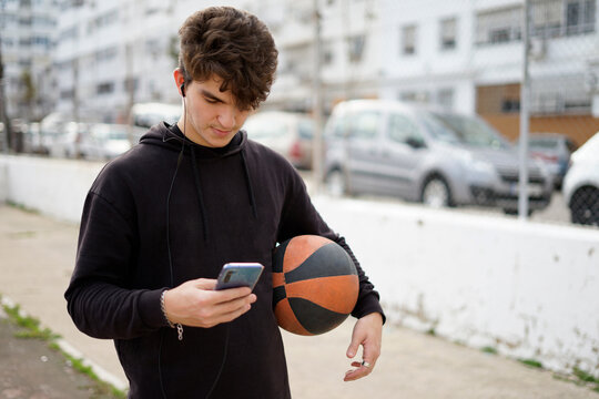 stock image of young man looking at cell phone with one hand and holding a basketball with the other on basketball court, horizontal image with copy space