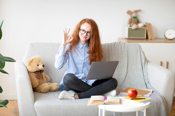 Girl in glasses using laptop for elearning, showing ok gesture