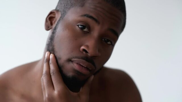 Man skincare. Male grooming. Morning routine. Confident satisfied cheerful handsome shirtless African guy touching face beard smiling isolated on light neutral background.