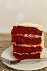 A slice of red velvet cake with cream cheese frosting on a plate with a fork and a festive cream backdrop 