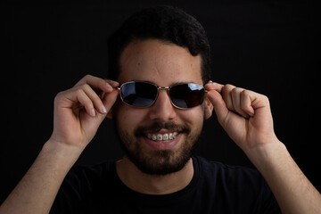 Obraz premium Young man frontal head shot with black background. He is smiling and taking off his sunglasses. He has a casual look with beard and t-shirt.